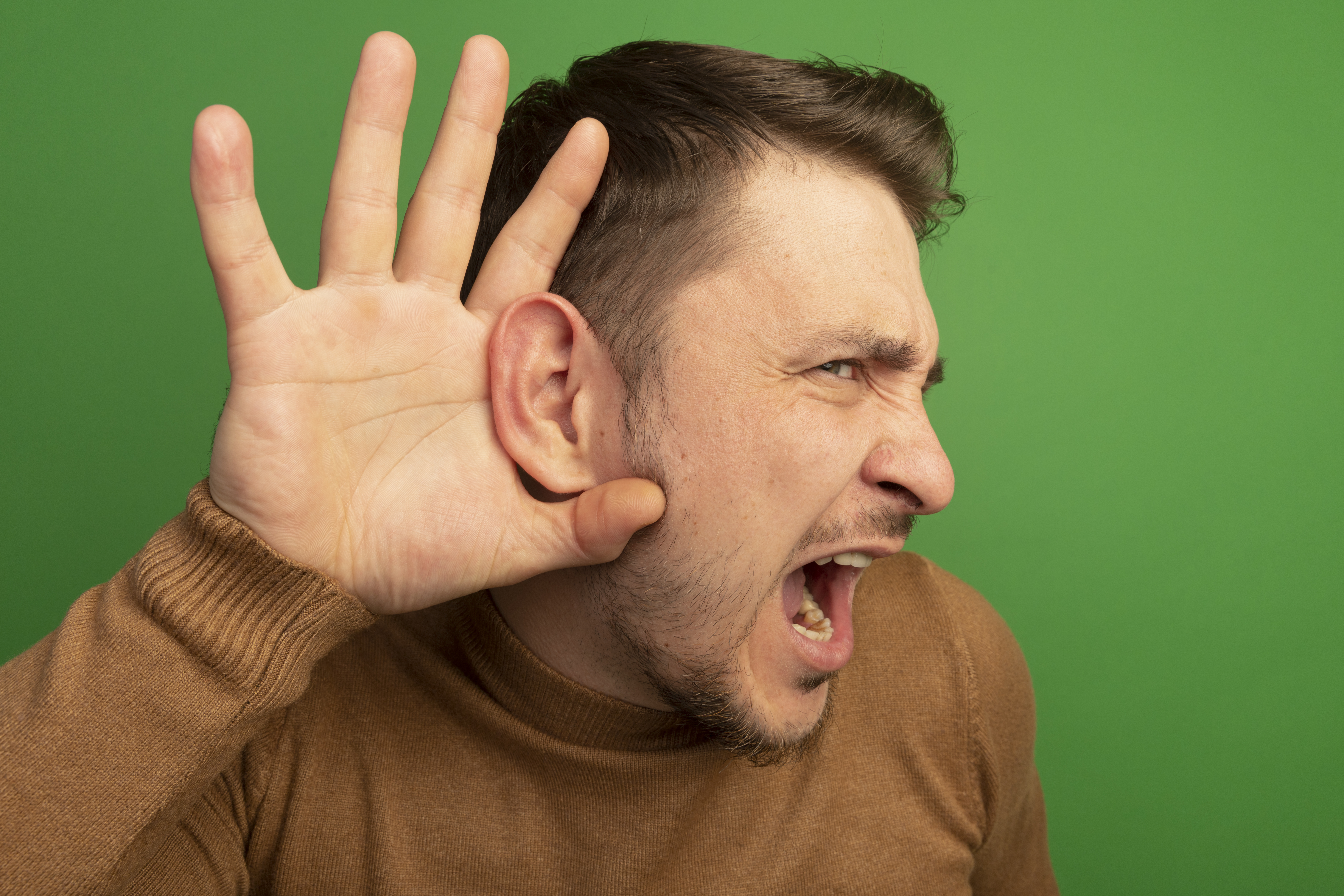 close-up view of curious young blonde handsome man keeping hand behind ear looking straight doing i can't hear you gesture isolated on green background